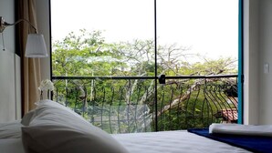 A hotel room with a large window, a bed with white bedding, a blue accent pillow, and a view of trees and a balcony railing. - Flor Inn Praia Hotel (Florianópolis)