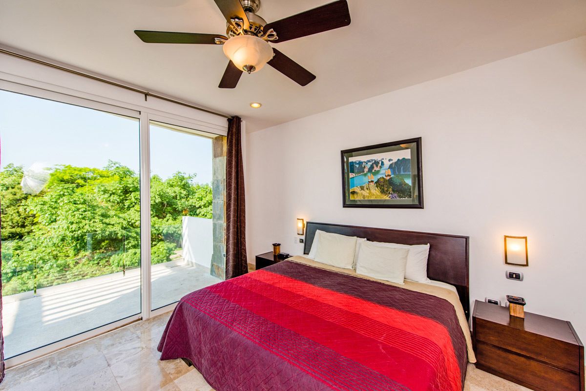 A bedroom with a large bed, a ceiling fan, a painting on the wall, and a view of greenery through the window.
