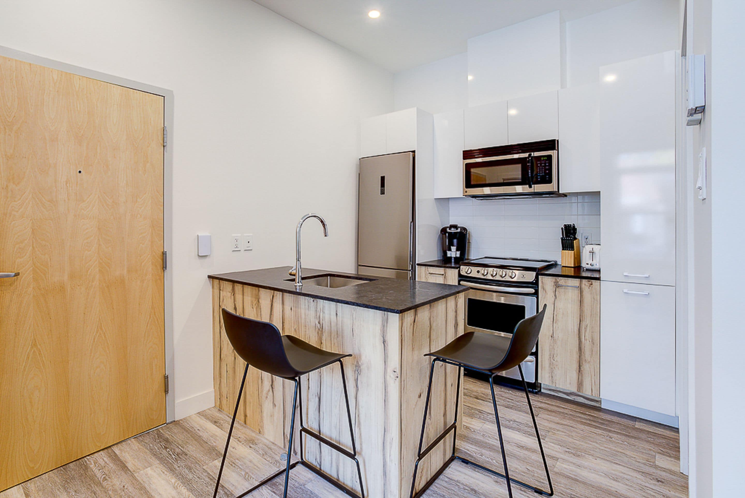 A compact kitchen with a wooden island, two bar stools, a stainless steel refrigerator, a microwave, and an oven.