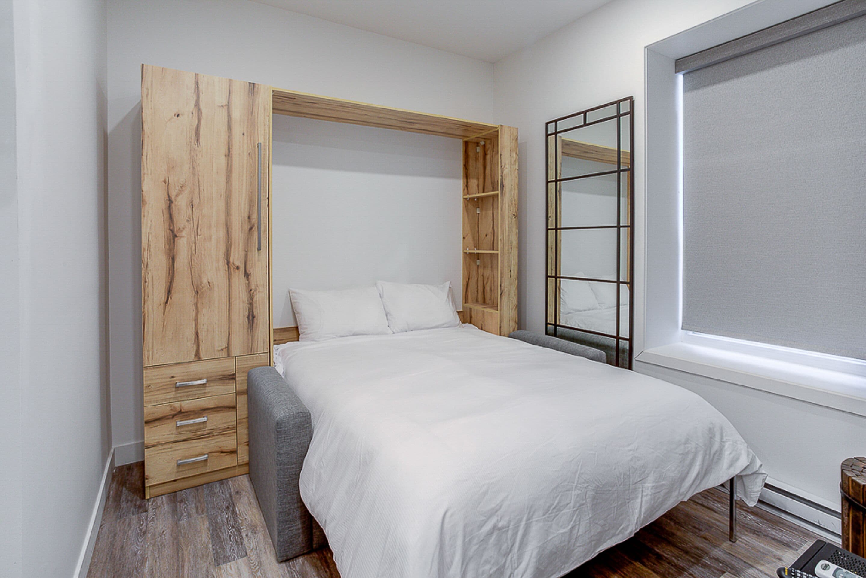 A bedroom with a large bed, a wooden wardrobe, a bench, and a window with blinds.