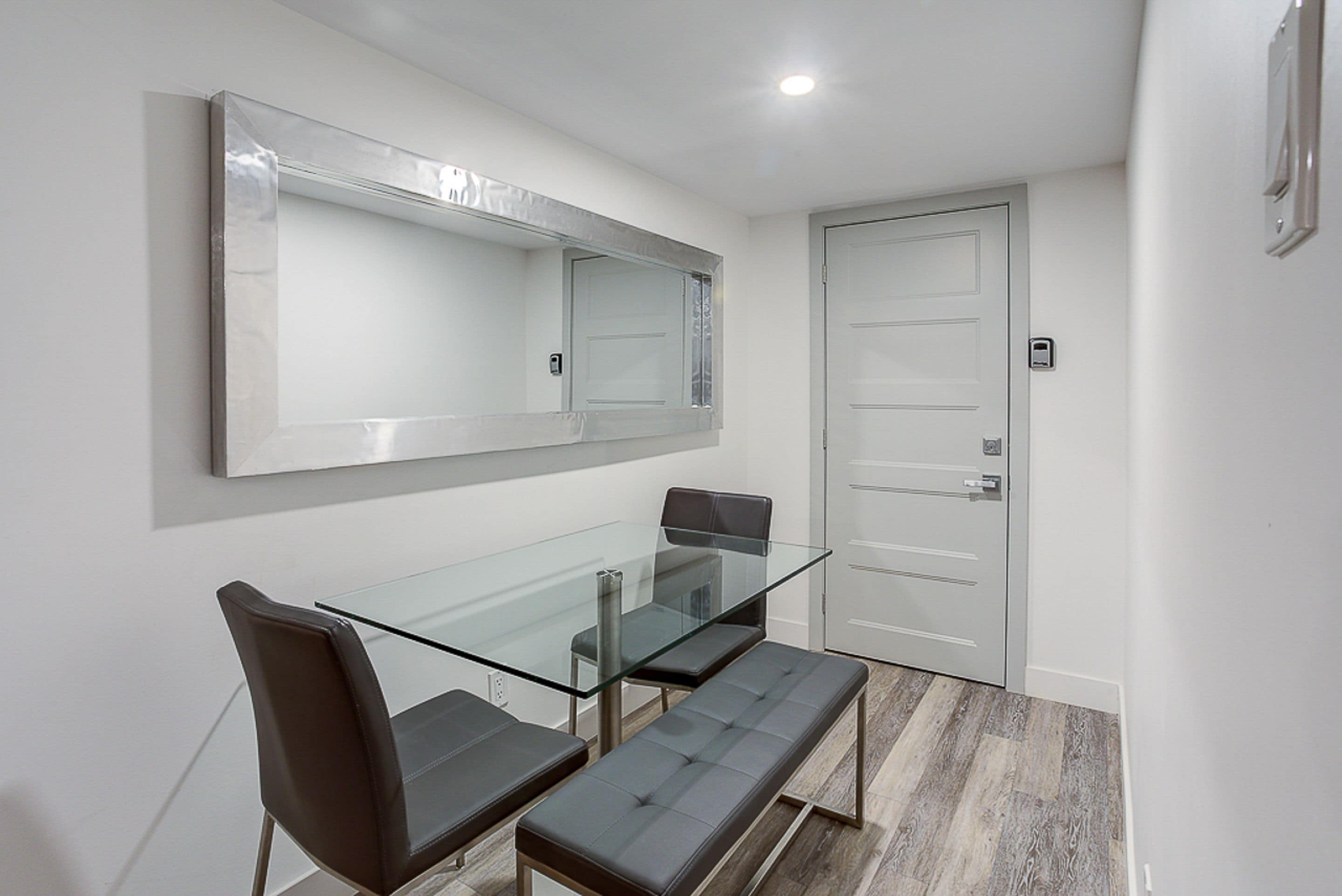 A modern conference room with a glass table, two leather chairs, a bench, and a large mirror on the wall.