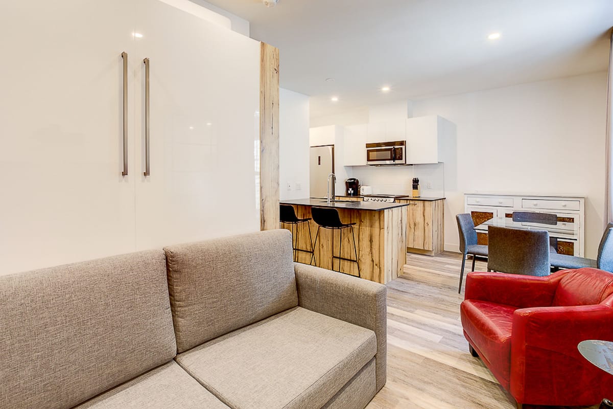 Un salon moderne avec un canapé, un fauteuil rouge et un îlot central en bois doté de tabourets de bar.