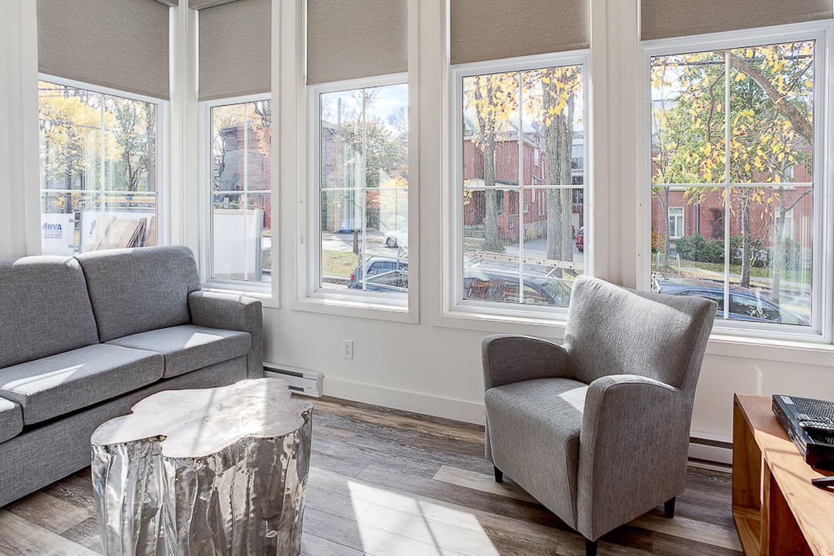 Un salon moderne avec un canapé gris, un fauteuil gris et une table basse en bois. De grandes fenêtres offrent une vue sur la rue et les arbres.