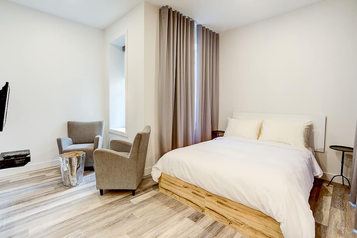Une chambre d’hôtel moderne avec un lit en bois, un fauteuil gris, une petite table d’appoint et une grande fenêtre avec des rideaux.