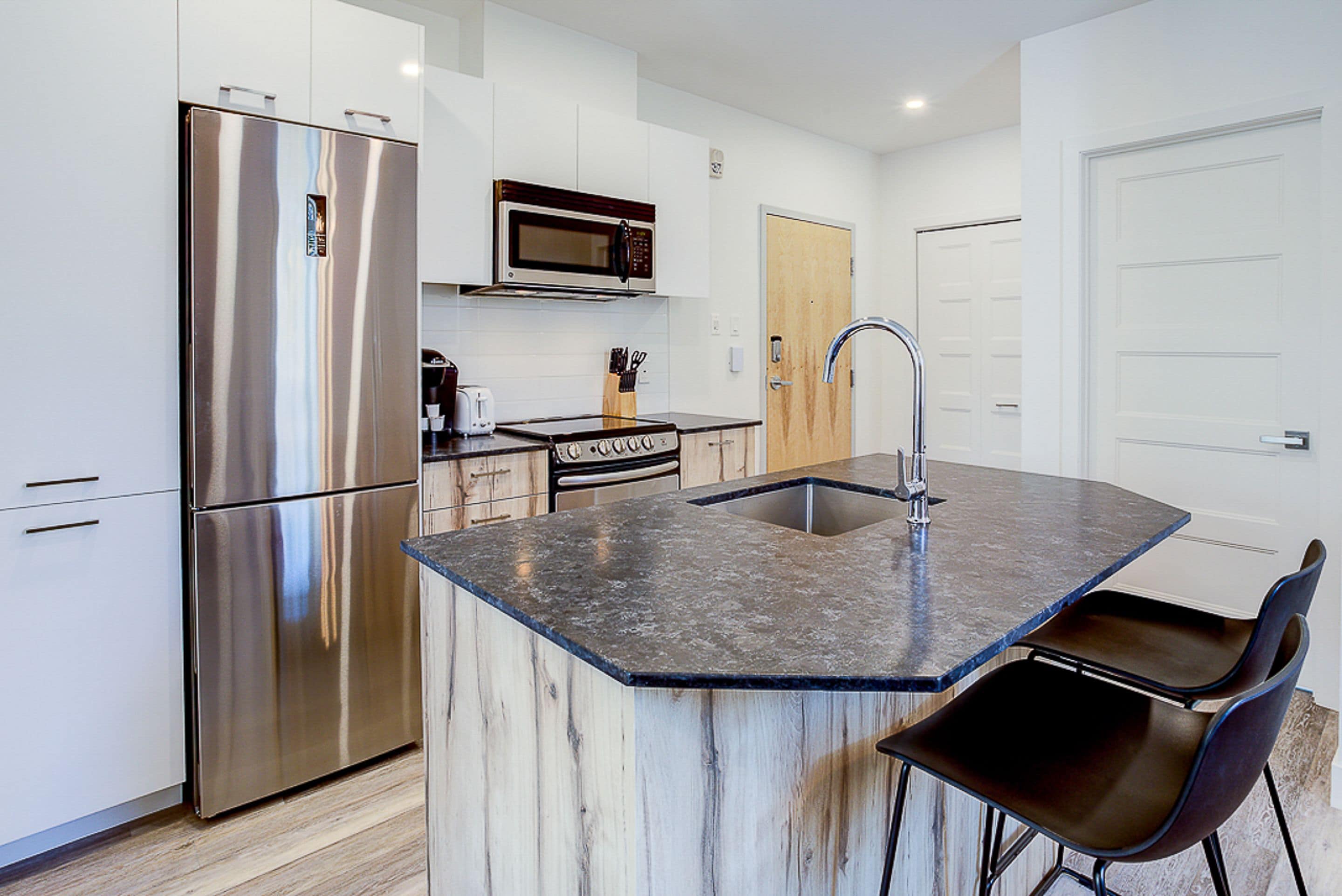 A modern kitchen with a stainless steel refrigerator, a microwave, and a sink with a faucet. There are two black chairs and a wooden island with a marble countertop.