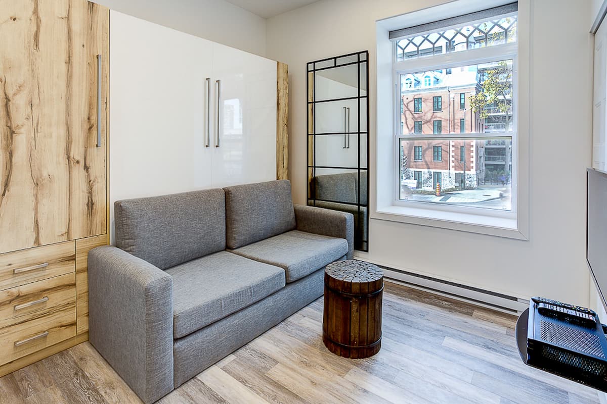 Un salon moderne avec un canapé gris, un meuble en bois et une grande fenêtre donnant sur des immeubles.