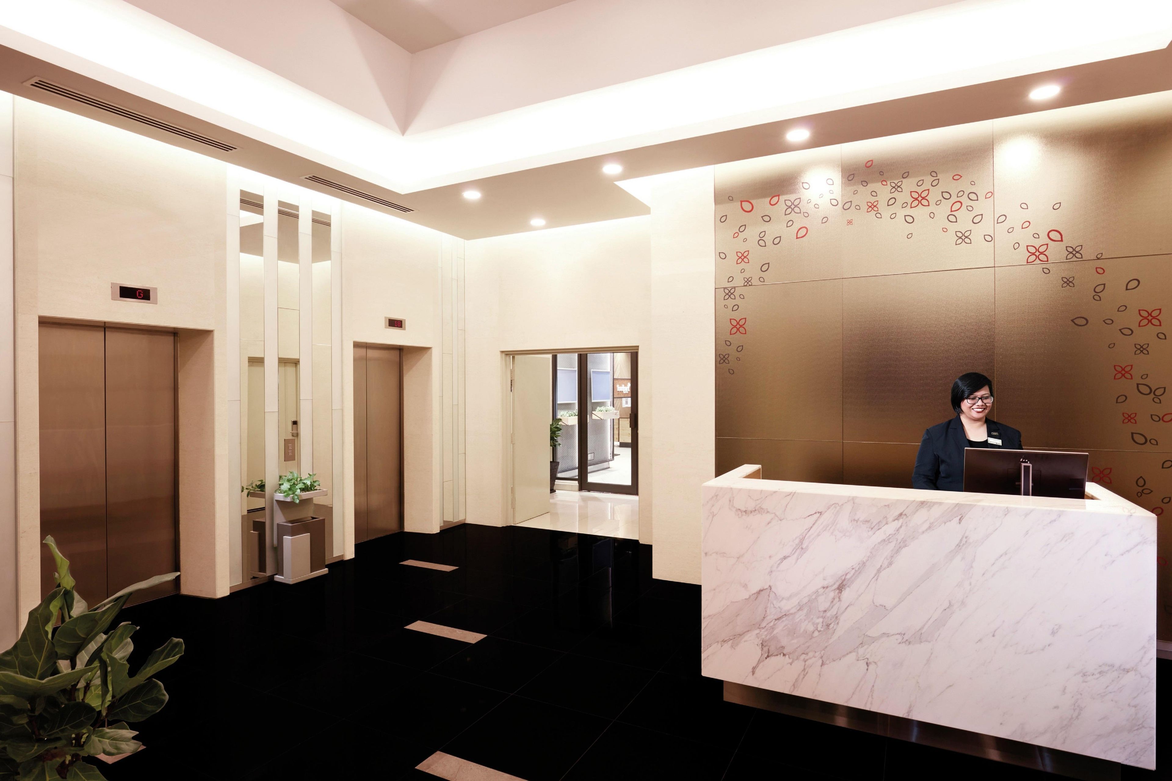 Zona de recepción de un hotel con mostrador de mármol, ascensores y una recepcionista.