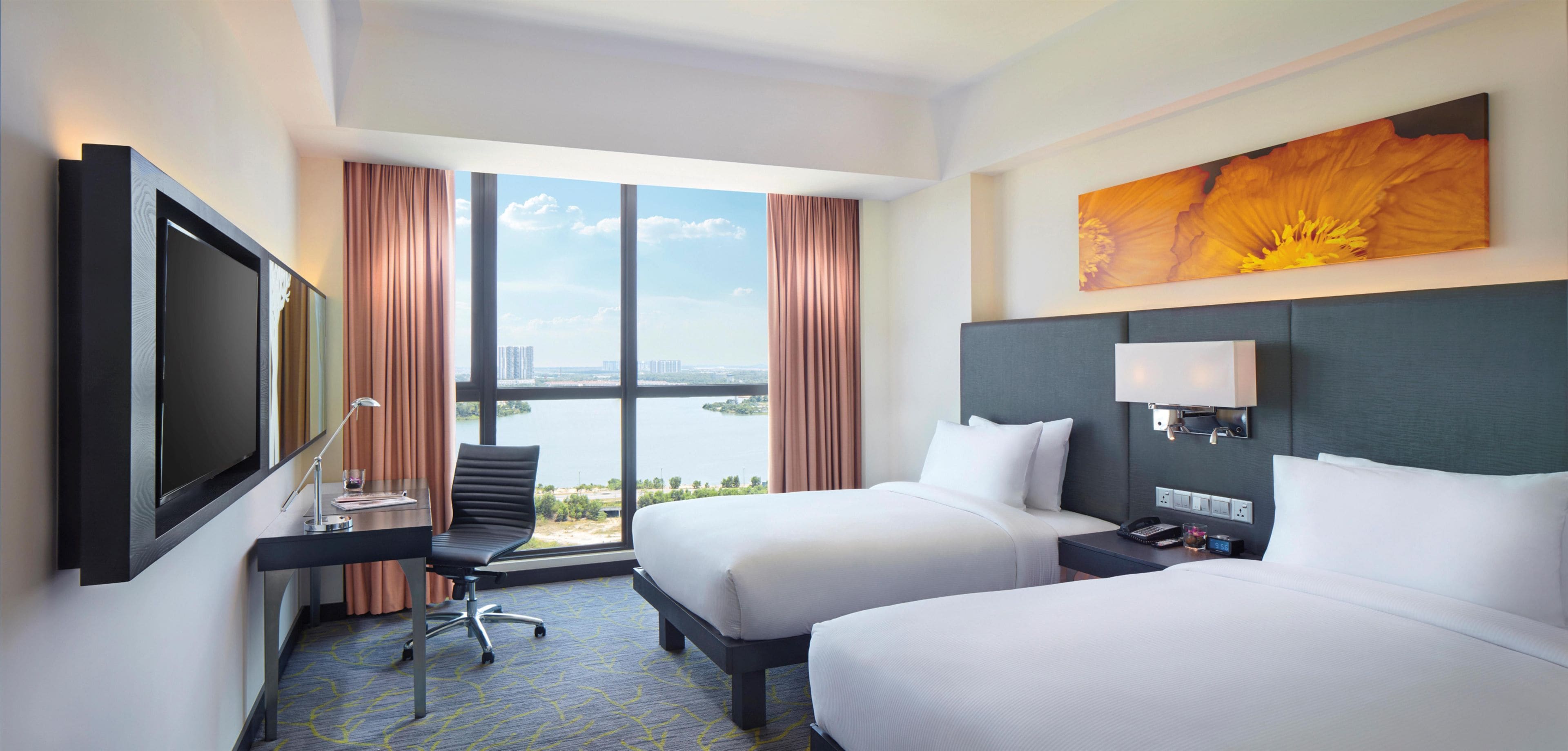 A hotel room with two beds, a desk with a chair, a TV, and a large window offering a view of a body of water and buildings.