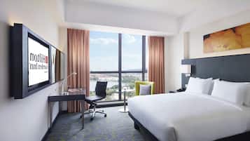 A hotel room with a large bed, a desk with a chair, a TV mounted on the wall, and a view of the city through large windows.