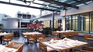 An outdoor dining area with wicker furniture and tables set for guests, a glass ceiling, and a view of the building's interior. - Hilton Garden Inn Puchong (Puchong)