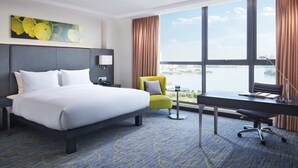 A hotel room with a large bed, a desk, a chair, and a view of a body of water through large windows. - Hilton Garden Inn Puchong (Puchong)