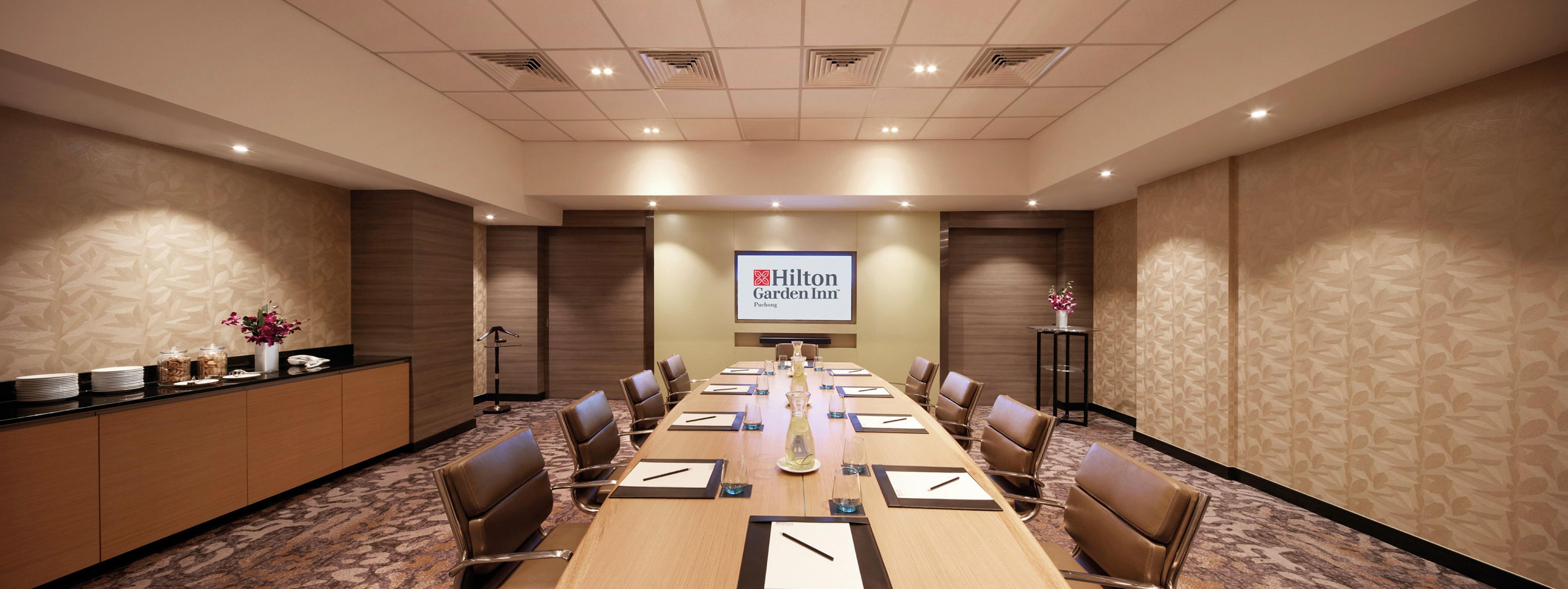 Un salón de conferencias con una larga mesa, sillas, un atril y una pantalla mostrando el logotipo de Hilton Garden Inn.