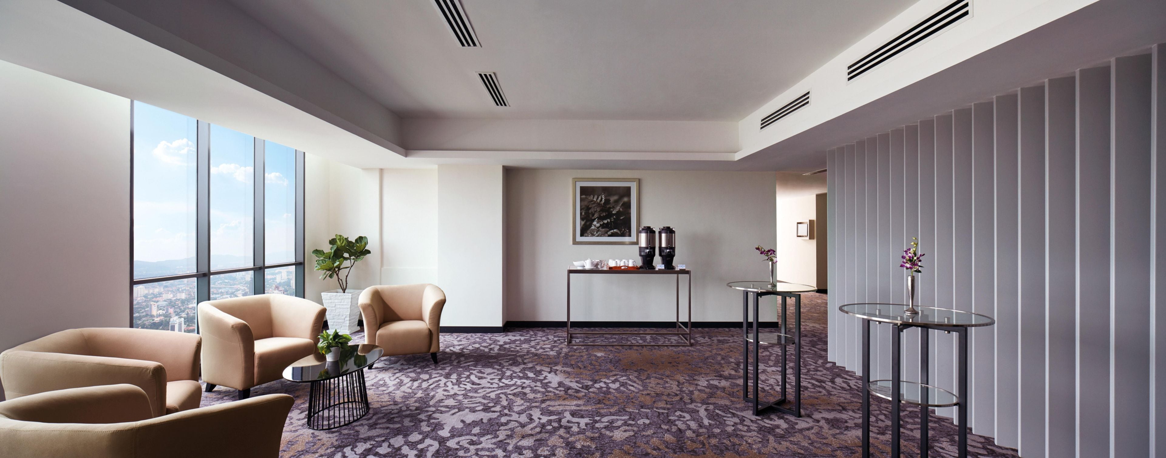 Una habitación de hotel moderna con ventanas amplias, suelo alfombrado, una zona de estar con sillas, una mesita y una planta decorativa.