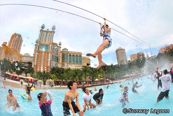 Parque acuático con tirolesa, piscinas y un paisaje urbano al fondo.