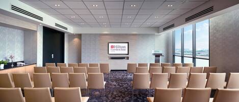 Un salón de conferencias con filas de sillas, un atril y una pantalla que muestra el logotipo de Hilton Garden Inn.