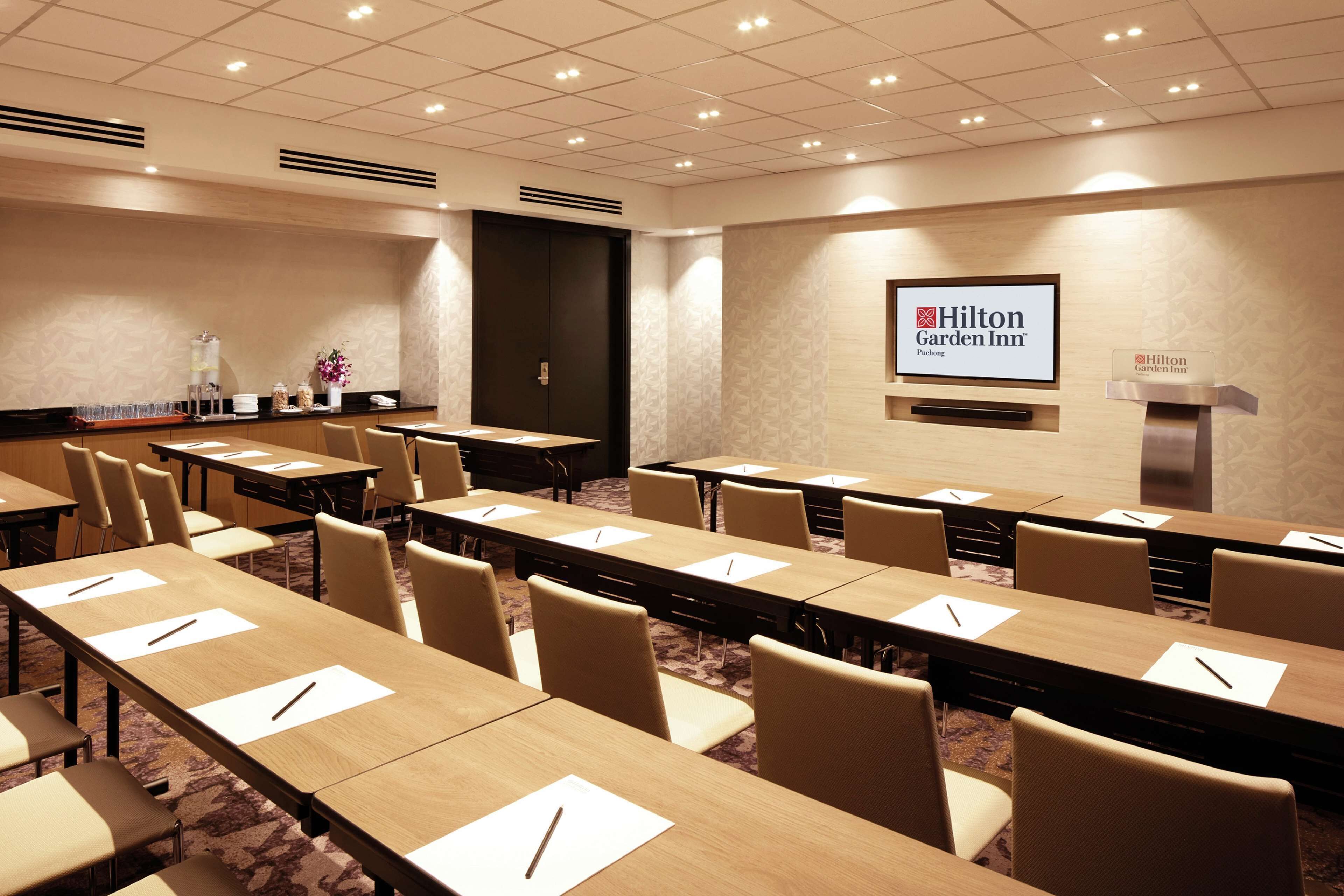 Un salón de conferencias con mesas largas, sillas y una pantalla mostrando el logotipo de Hilton Garden Inn.