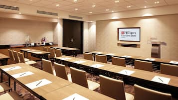 Un salón de conferencias con mesas largas, sillas y una pantalla mostrando el logotipo de Hilton Garden Inn.
