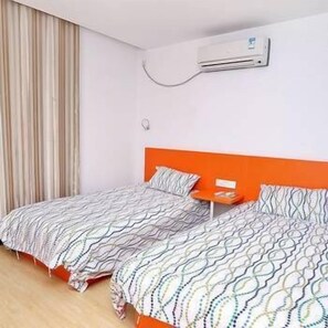 A hotel room with two beds, an orange headboard, a bedside table with a lamp, and an air conditioning unit on the wall.