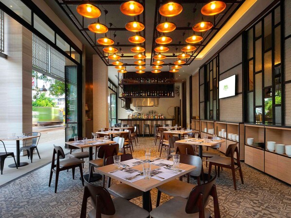 A restaurant interior with multiple tables, chairs, hanging pendant lights, a bar area, and large windows.