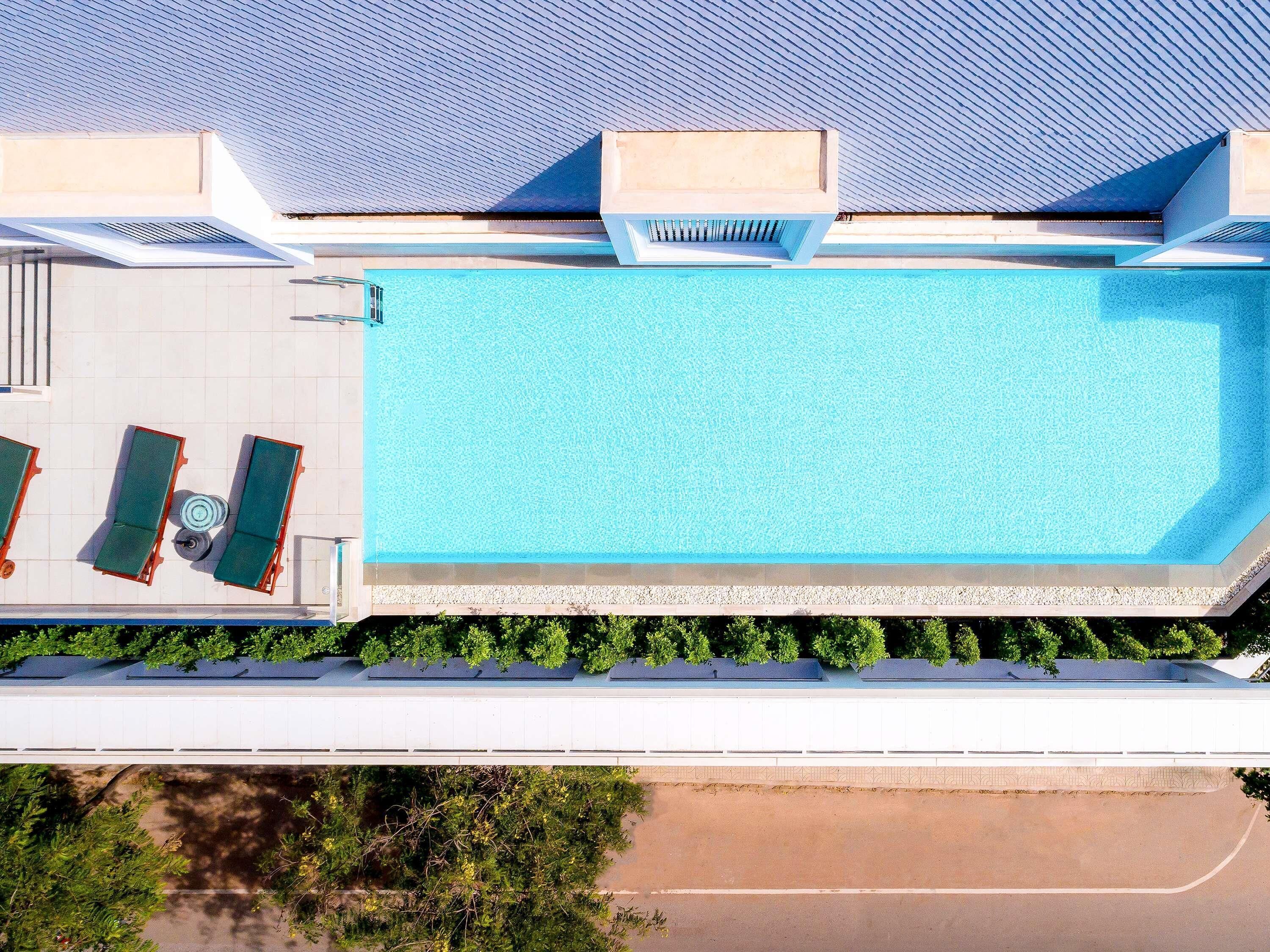 An aerial view of a swimming pool with two sun loungers and a small table, surrounded by greenery and a covered roof structure.