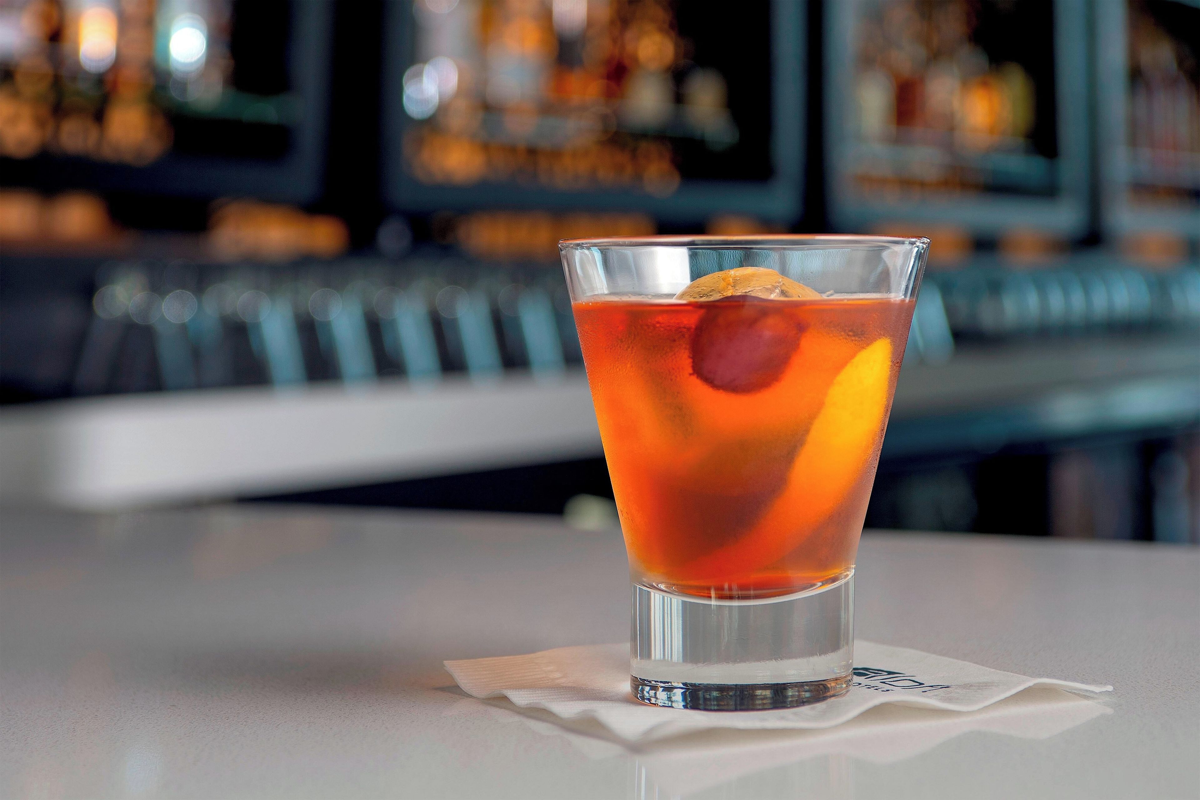 Un vaso de bebida color ámbar con un cubito de hielo y una rodaja de naranja, sobre un servilleta blanca en un bar con fondo desenfocado.