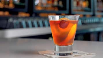 Un vaso de bebida color ámbar con un cubito de hielo y una rodaja de naranja, sobre un servilleta blanca en un bar con fondo desenfocado.