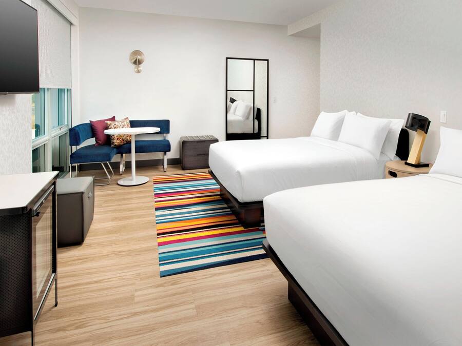 Habitación de hotel con dos camas, un escritorio, una silla, una mesita, un espejo y un colorido tapete a rayas.