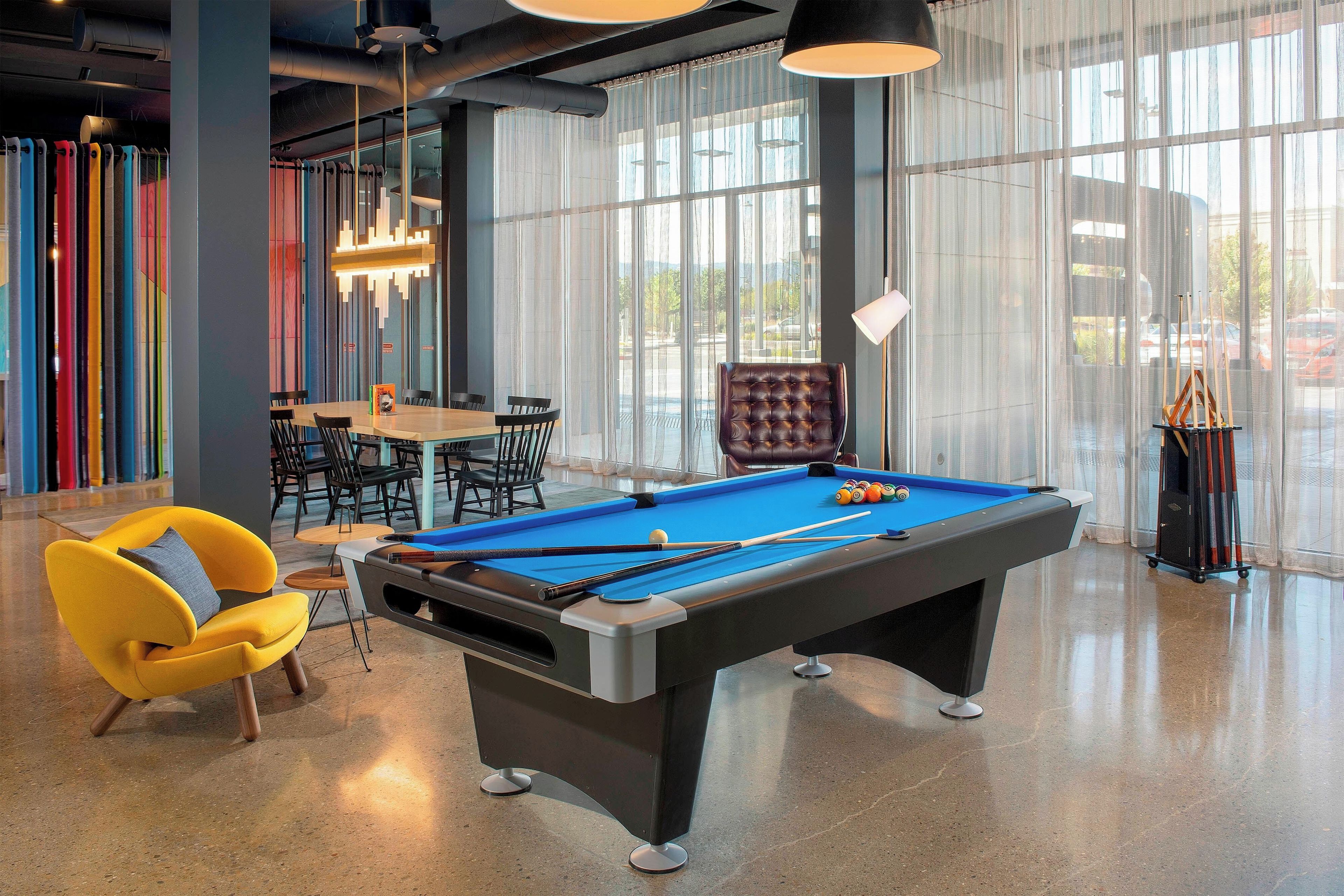 A pool table in a well-lit room with large windows, a yellow armchair, and a billiards rack.
