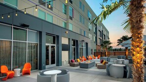 Un área de estar al aire libre con mobiliario moderno, una palmera y un edificio de grandes ventanales y techo plano.