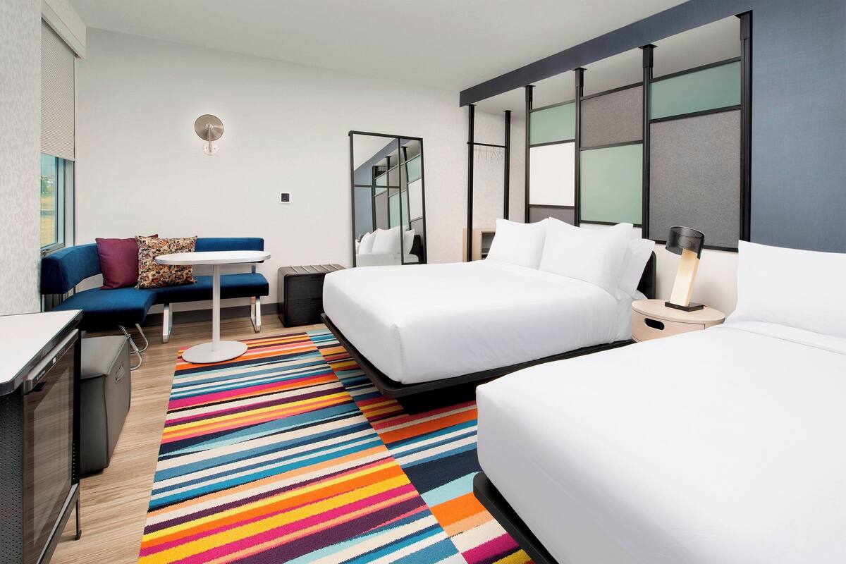 Quarto de hotel moderno com uma cama grande, um sofá, uma escrivaninha e um tapete colorido listrado.