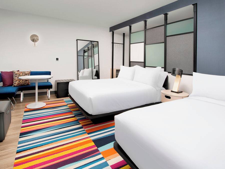 A modern hotel room with a large bed, a sofa, a desk, and a colorful striped rug.