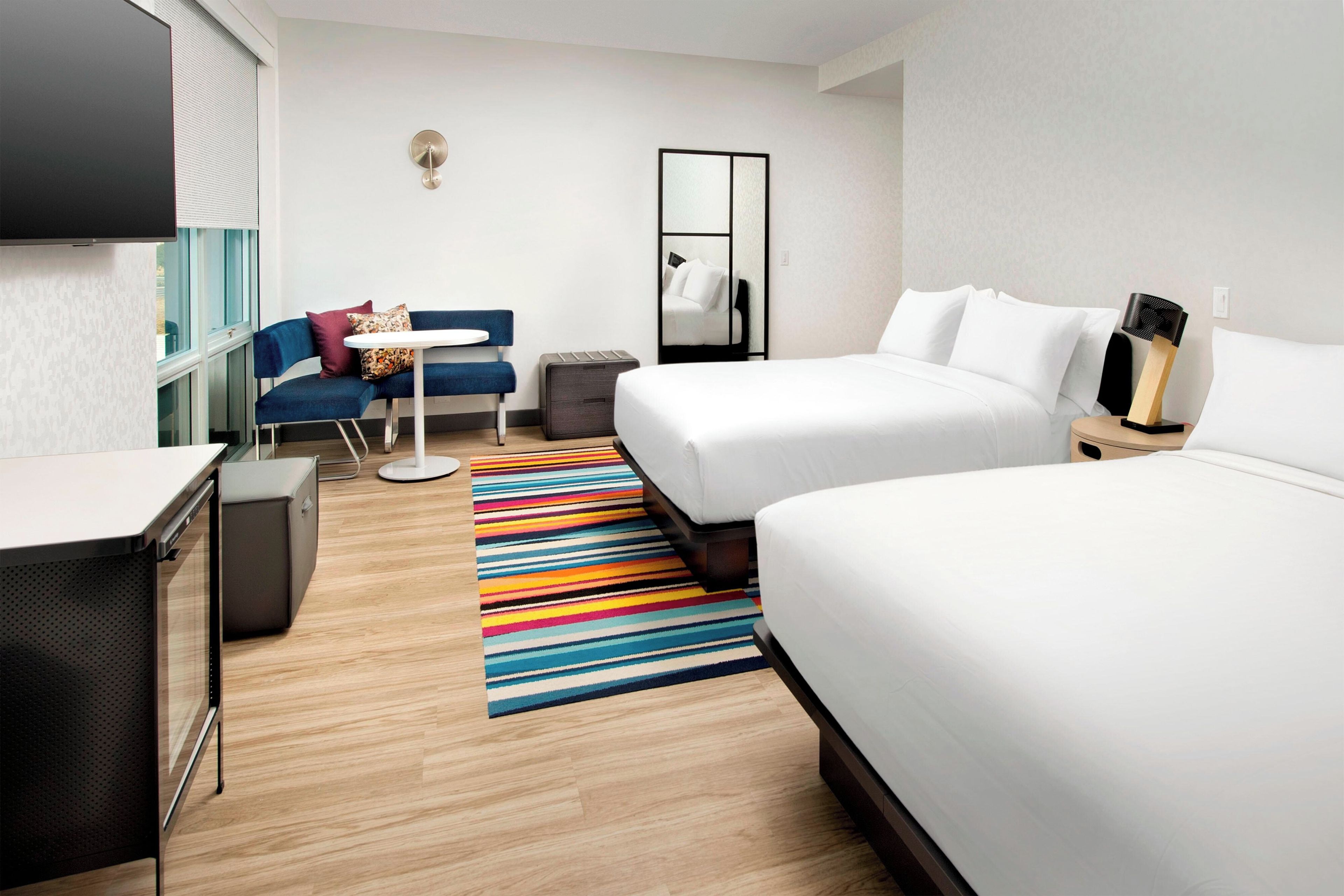A hotel room with two beds, a desk, a chair, a small table, a colorful striped rug, and a mirror.
