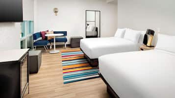 Une chambre d’hôtel avec deux lits, un bureau, une chaise, une petite table, un tapis rayé aux couleurs vives et un miroir.
