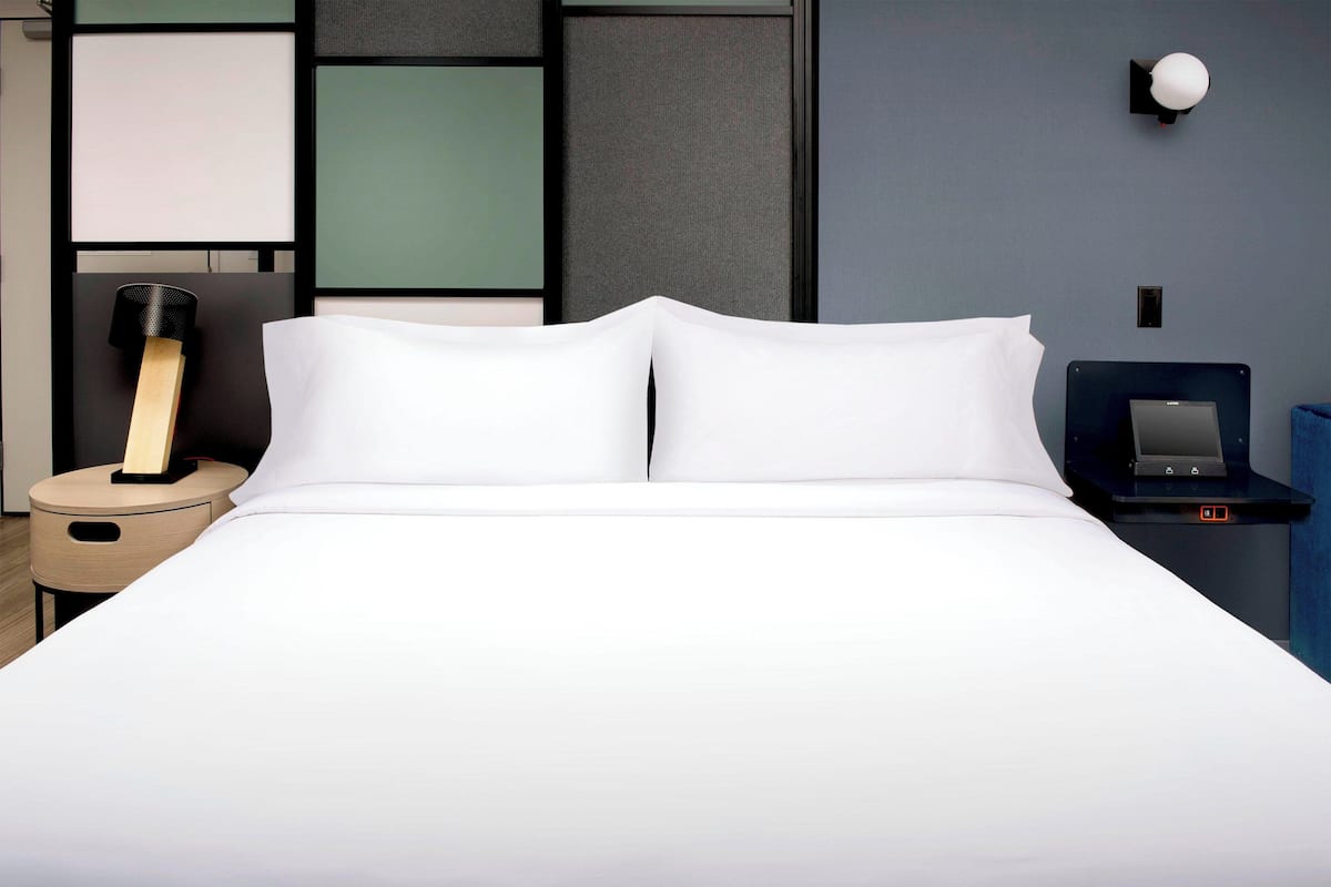Cama arrumada em quarto de hotel, com mesa de cabeceira, abajur e relógio digital.