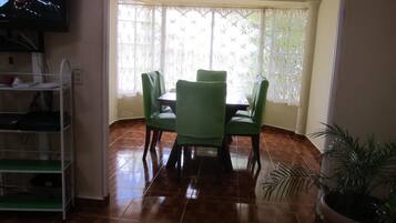 Uma área de refeições com mesa e cadeiras, uma TV na parede e um vaso com planta.
