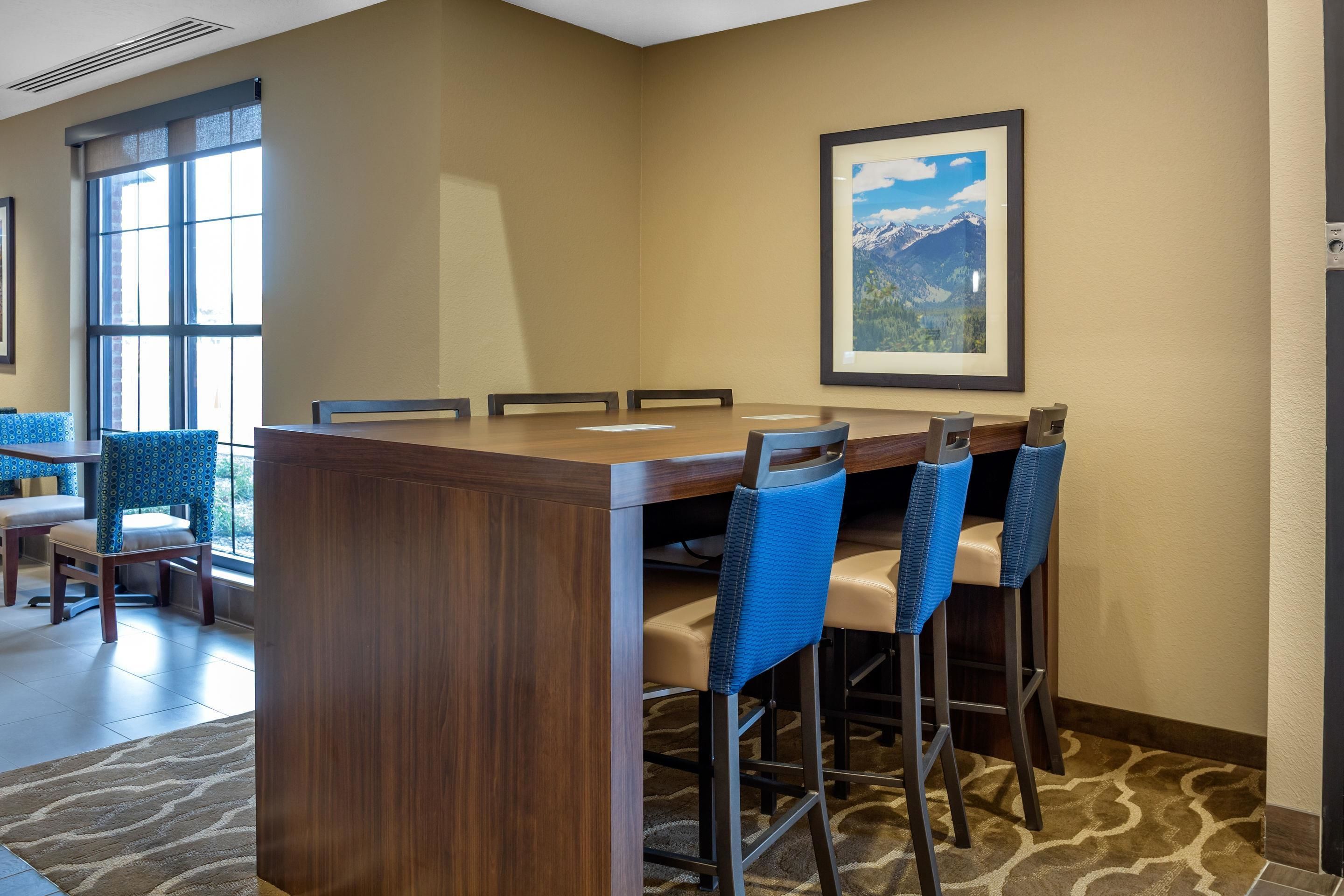 A hotel bar with a wooden bar counter, blue upholstered bar stools, and a framed picture of a mountain landscape on the wall.