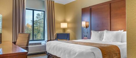 A hotel room with a large bed, a desk, a chair, a window with curtains, and a wall-mounted lamp.