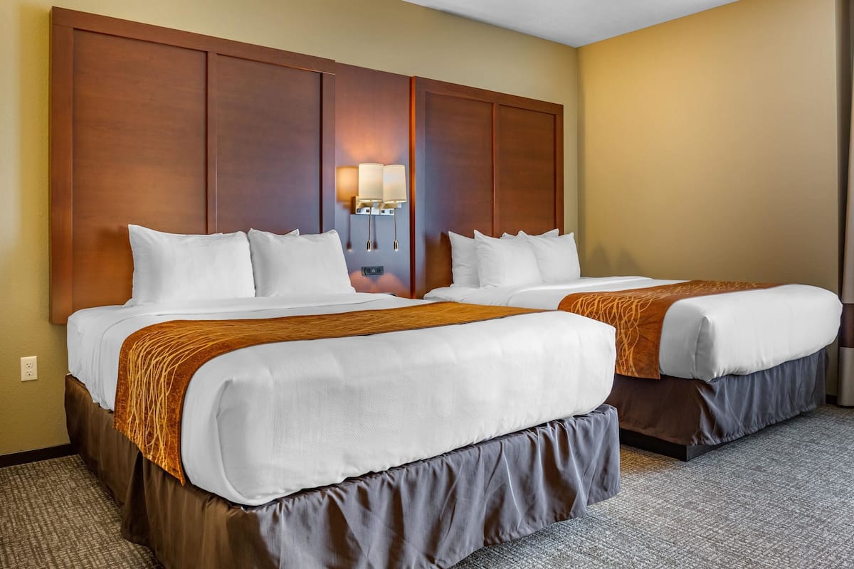 Habitación de hotel con dos camas, cabecera de madera, mesita de noche con lámpara y suelo alfombrado.