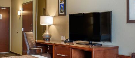 A hotel room with a bed, a desk with a television, a lamp, a chair, and a framed picture on the wall.