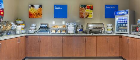 Zona de desayuno tipo buffet con diversas opciones de comida, una máquina de café y un refrigerador.