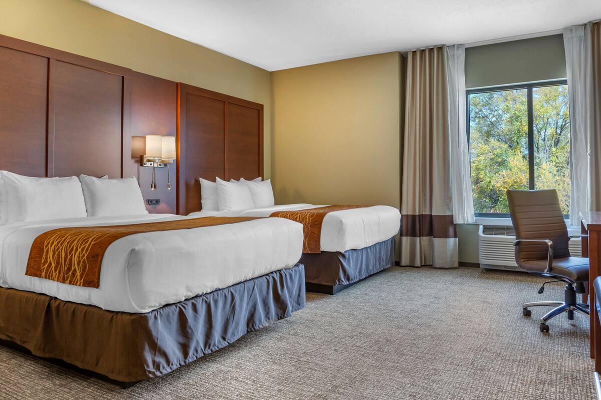 Habitación de hotel con dos camas, un escritorio, una silla y un amplio ventanal con cortinas.