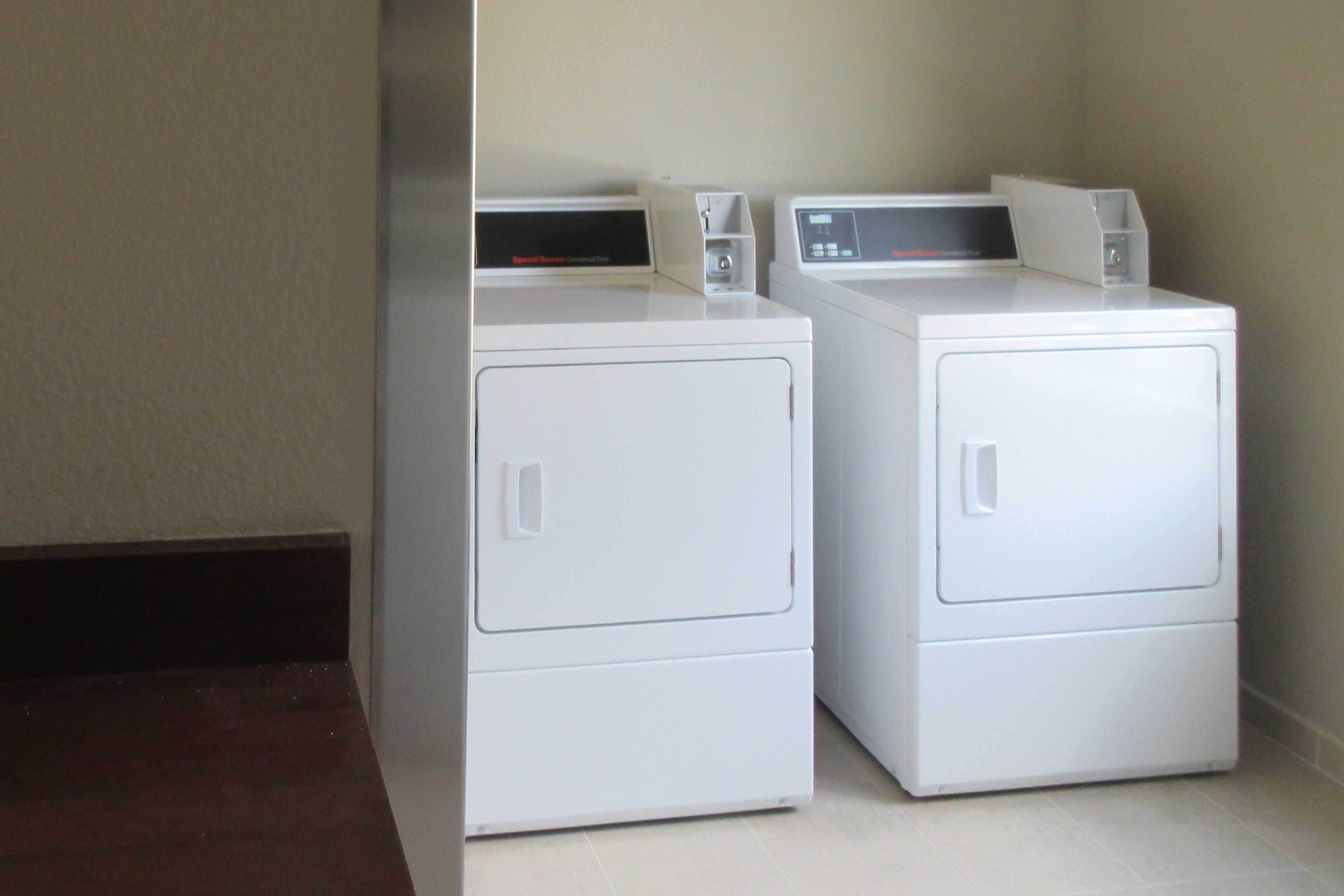 Two white front-loading washers in a corner with a dark floor and light walls.