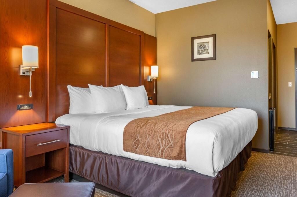 A hotel room with a large bed, wooden headboard, two bedside lamps, a nightstand, and a framed picture on the wall.