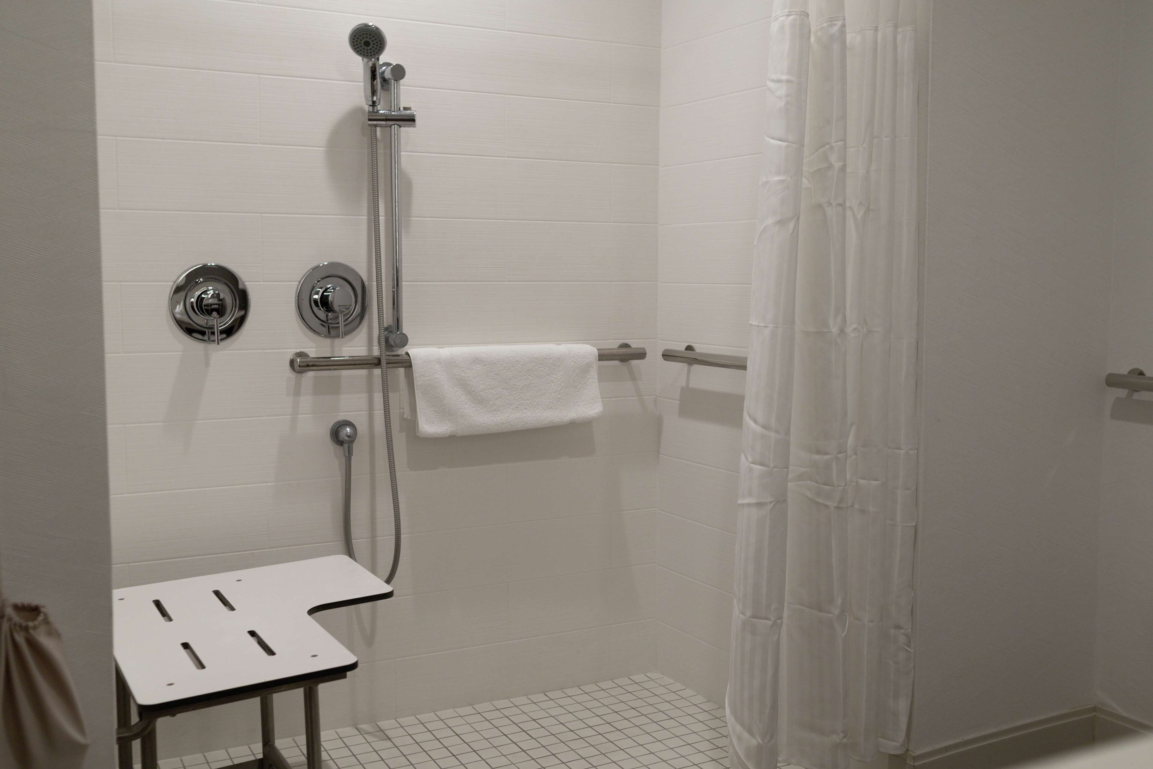 A shower area with a white tiled wall, a handheld showerhead, a towel rack, and a small table.