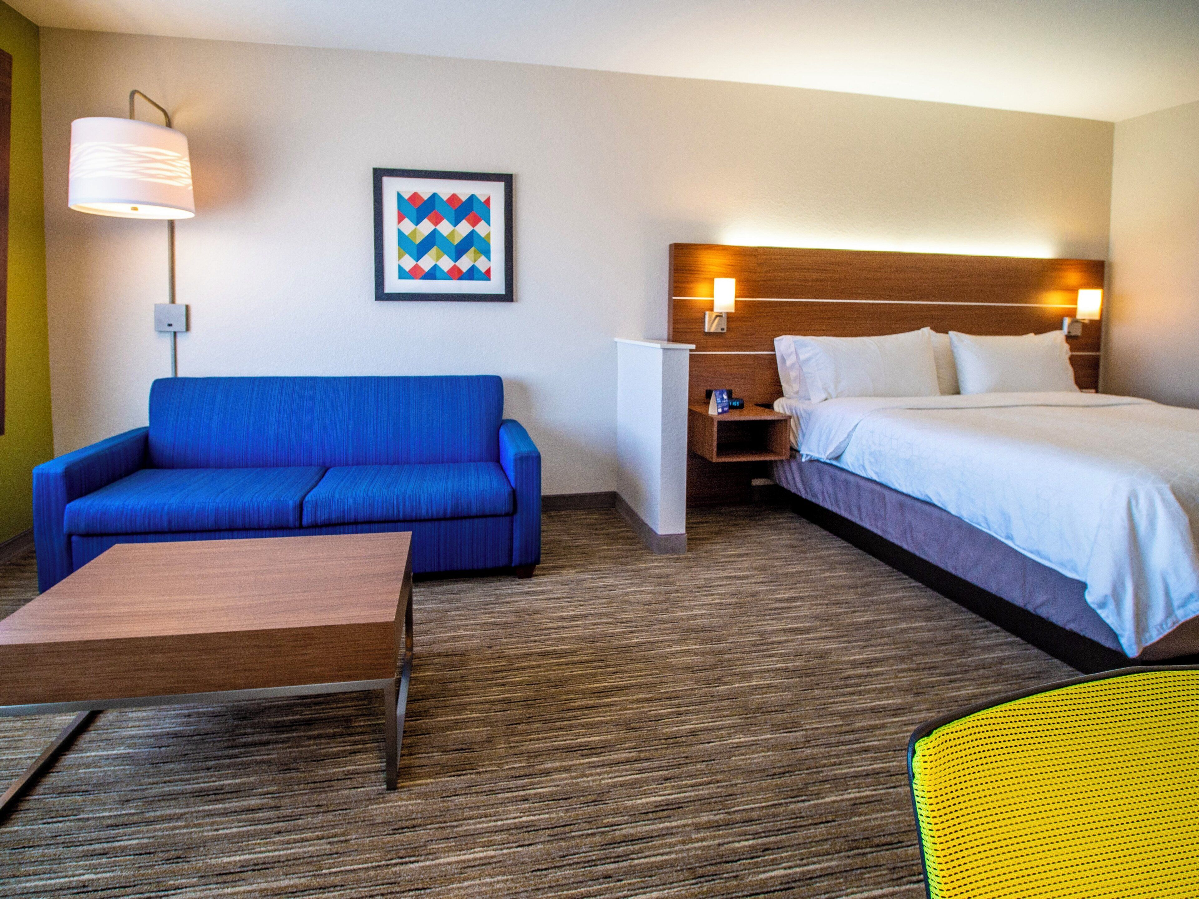 A hotel room with a blue sofa, a wooden table, a bed with white linens, a nightstand, a wall-mounted lamp, and a framed geometric artwork.