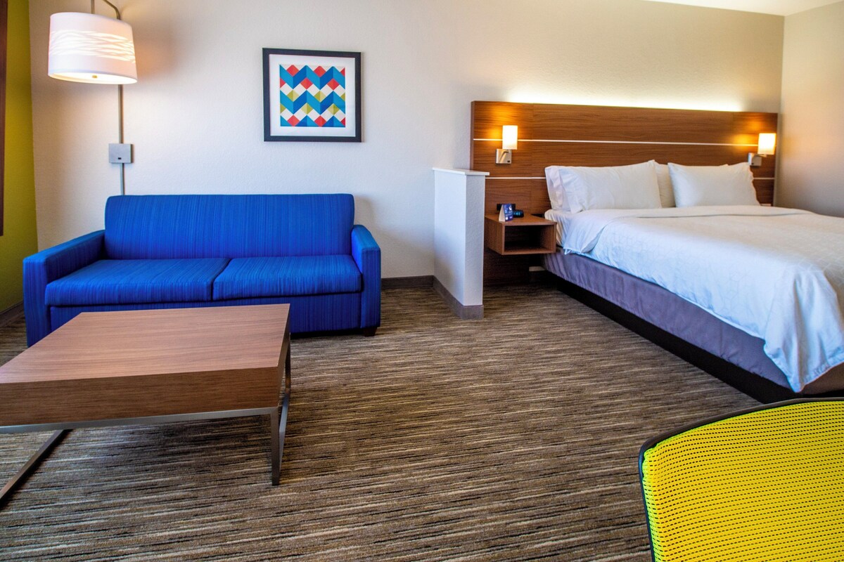 Quarto de hotel com um sofá azul, uma mesa de madeira, uma cama com roupa branca, uma mesinha de cabeceira, uma luminária na parede e uma obra de arte geométrica emoldurada.