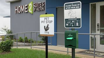 A hotel entrance with a 'Home 2 Suites by Hilton' sign, a pet pit stop sign, and a green waste disposal box.