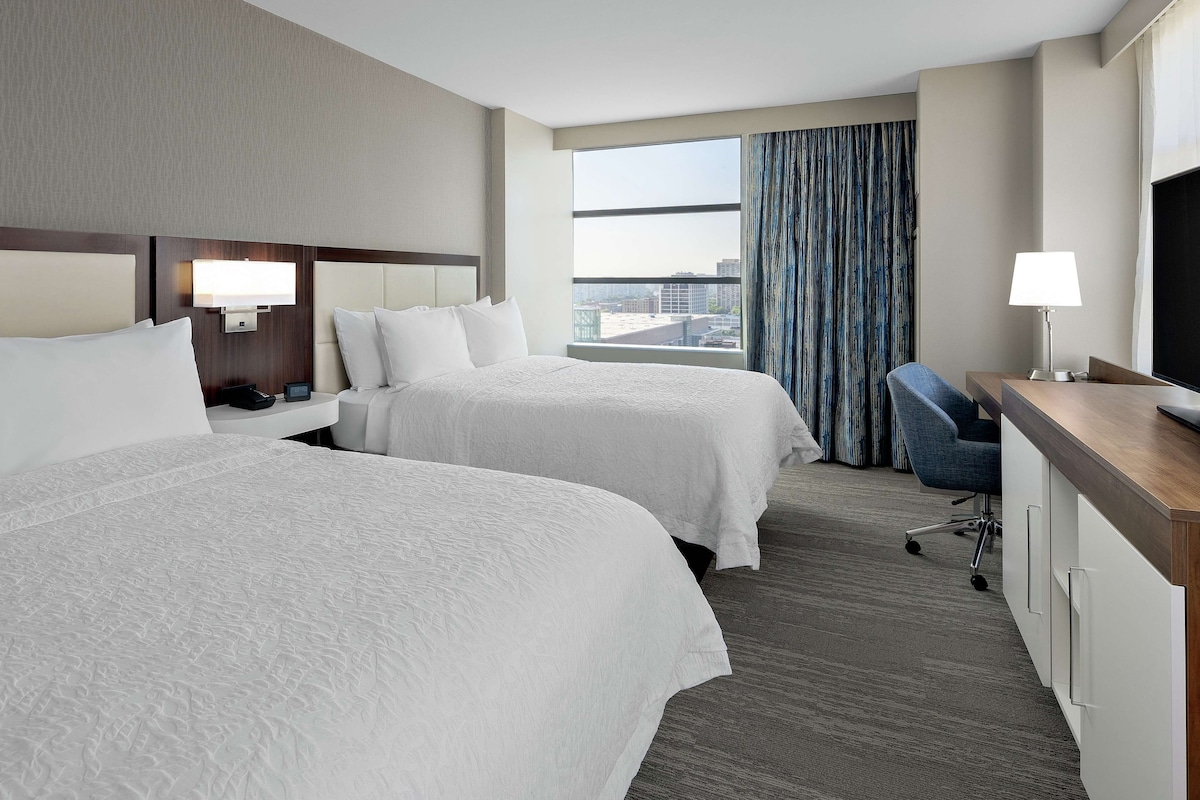 A hotel room with two beds, a desk, a chair, a TV, and a window with curtains.