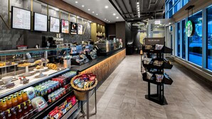 Interior de cafeteria moderna com balcão exibindo doces, bebidas variadas e petiscos.
