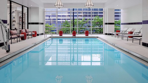 A modern indoor swimming pool with clear water, surrounded by lounge chairs and a view of buildings through large windows.